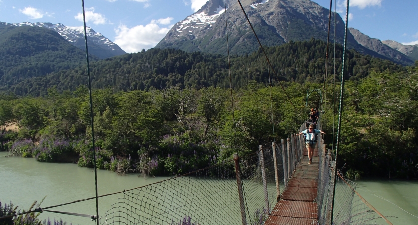 A group of people make their way over a swing bridge that hangs over a river. In the background, there are thick green trees in front of a mountain. 